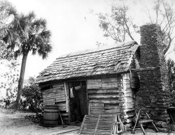 Man standing in the doorway of a log cabin near Turkey Creek - Tillman, Florida.